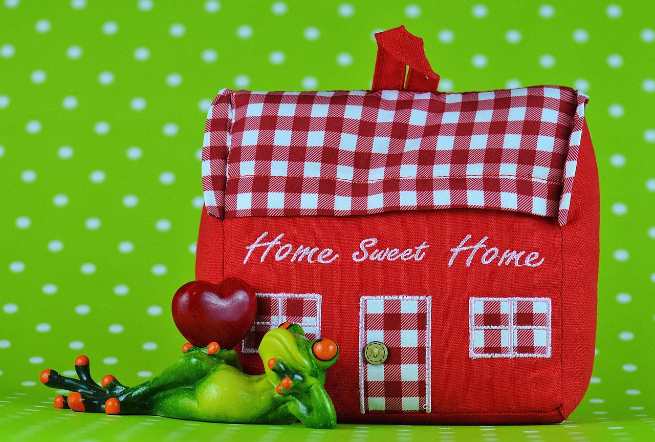 a frog figurine holding a red heart, lies by a 'home sweet home' red-checkered plush house in front of a green polka dot background
